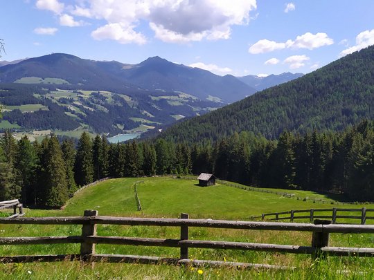 Your Plan de Corones/Kronplatz hotel on the slopes View of green meadows, wooden fence, forested mountains, and blue sky with clouds