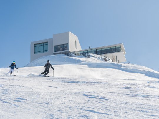 Your Plan de Corones/Kronplatz hotel on the slopes Two skiers skiing on snowy slope in front of modern building under clear sky