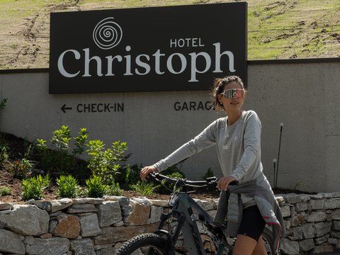 Willkommen in Olang: Hotel Christoph Frau mit Fahrrad vor dem Hotel Christoph Schild und Steinmauer