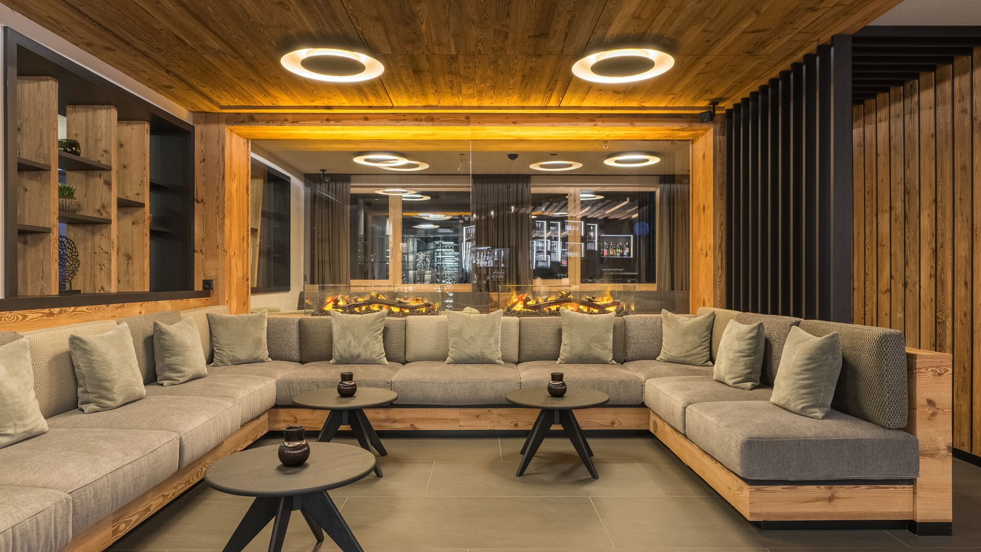 Willkommen in Olang: Hotel Christoph Moderner Aufenthaltsraum mit grauem Ecksofa und Kamin mit Holzinterieur