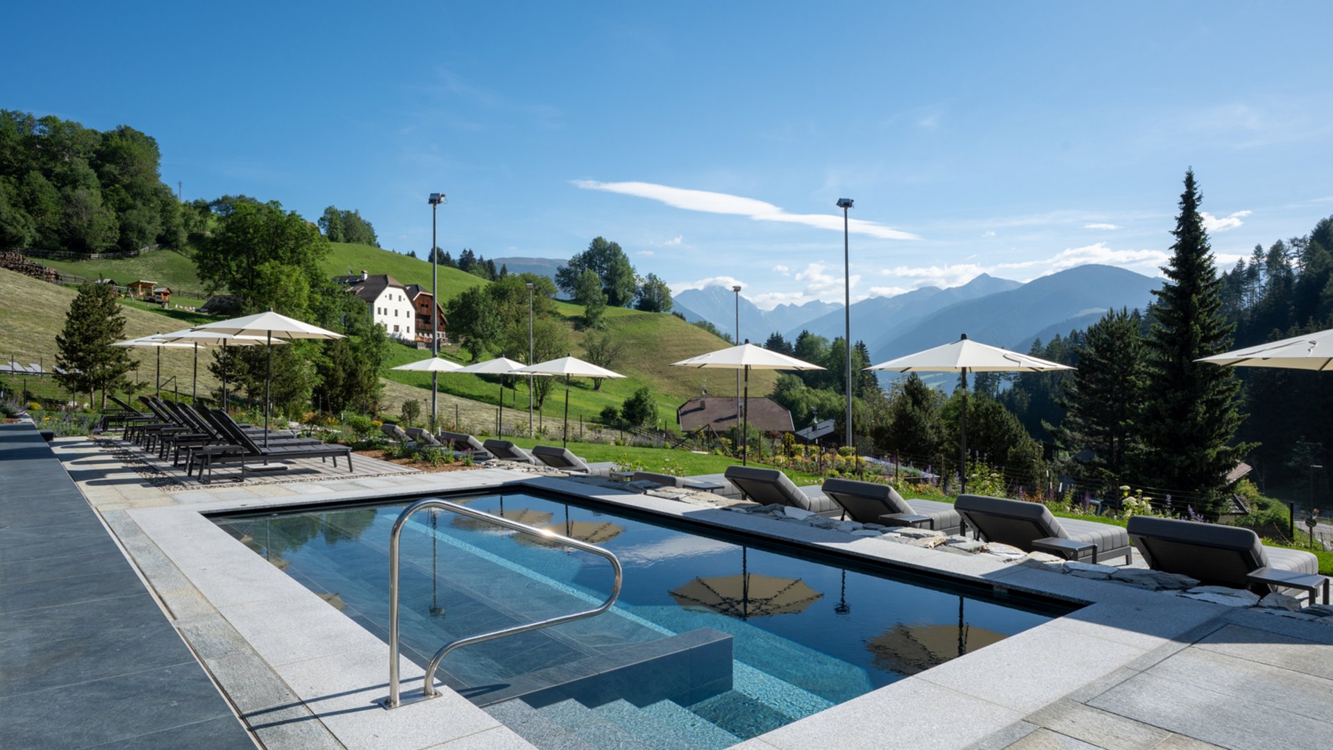 Willkommen in Olang: Hotel Christoph Pool mit Liegestühlen und Sonnenschirmen vor Berglandschaft bei klarem Himmel