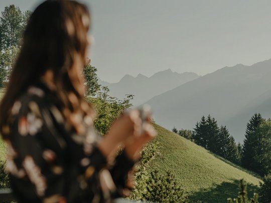 Your Plan de Corones/Kronplatz hotel on the slopes Woman with cup on balcony looking at green hills and mountains in background